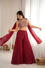 A woman modeling a deep red traditional outfit consisting of a heavily embroidered sleeveless crop top, matching wide-leg flared pants, and a flowing sheer dupatta, set against a bright indoor background with white curtains.