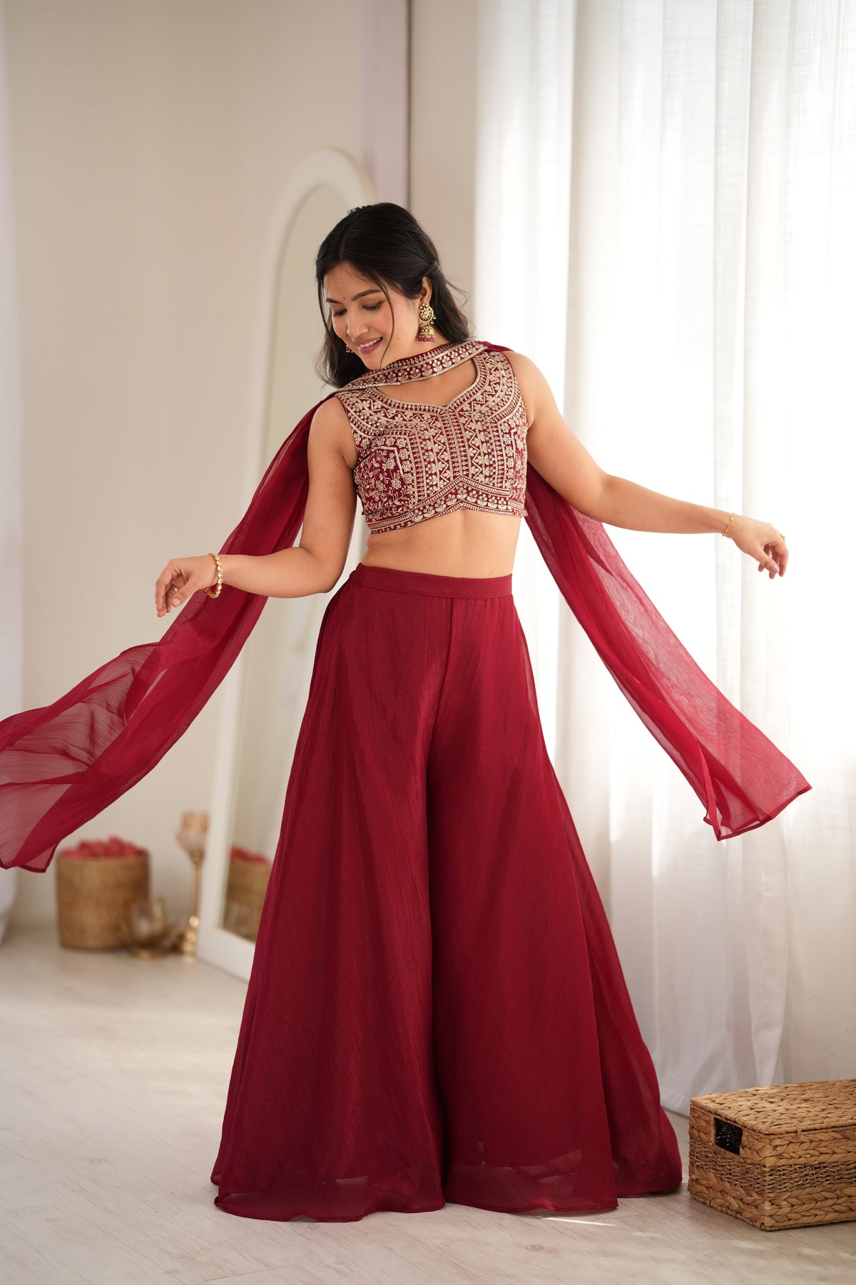 A woman modeling a deep red traditional outfit consisting of a heavily embroidered sleeveless crop top, matching wide-leg flared pants, and a flowing sheer dupatta, set against a bright indoor background with white curtains.