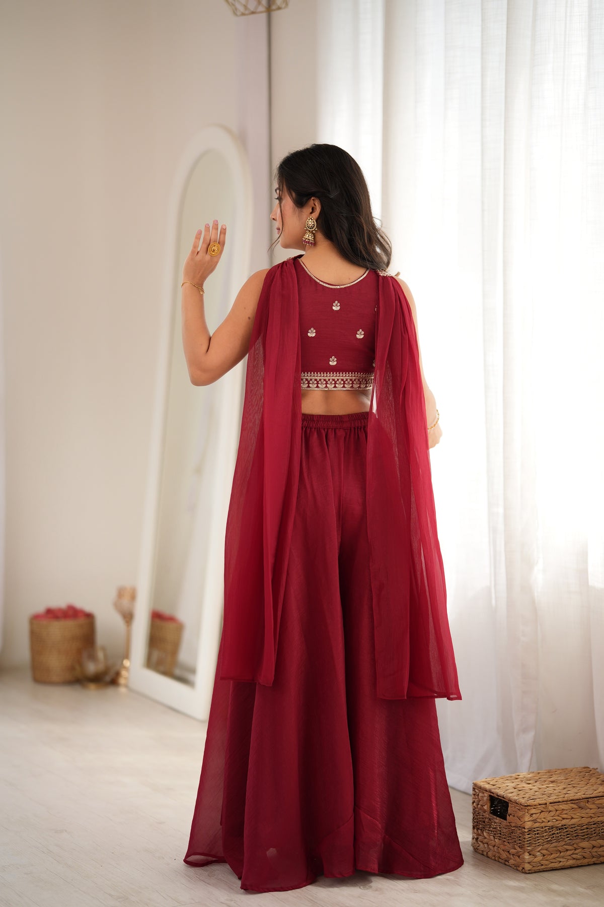 A woman stands in a bright indoor room wearing a deep maroon traditional outfit with a sleeveless crop top, wide-leg pants, and a matching sheer draped overlay. She is seen from the back, facing a mirror, with white curtains and neutral decor in the background.