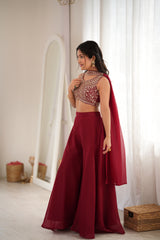 A woman models a traditional Indian outfit consisting of a deep red flared skirt, a heavily embroidered red and gold sleeveless blouse, and a matching red dupatta, set against a bright, minimalist indoor background with a mirror and sheer curtains.