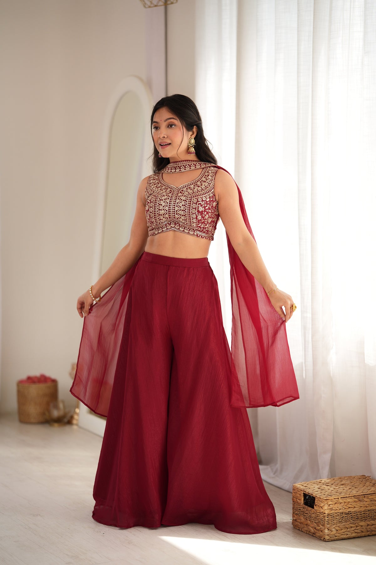 A woman modeling a traditional Indian outfit consisting of a deep red sleeveless crop top with intricate gold embroidery, matching wide-leg pants, and a sheer red dupatta, set against a bright, sunlit indoor background with white curtains.