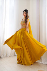 Woman in a yellow traditional outfit with a floral top and flowing skirt, standing against white curtains.