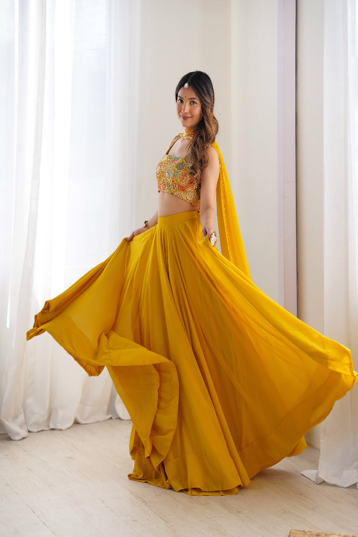 Woman in a yellow traditional outfit with a floral top and flowing skirt, standing against white curtains.