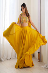 Woman in a yellow traditional outfit with a floral top and flowing skirt, standing against a white curtain background.
