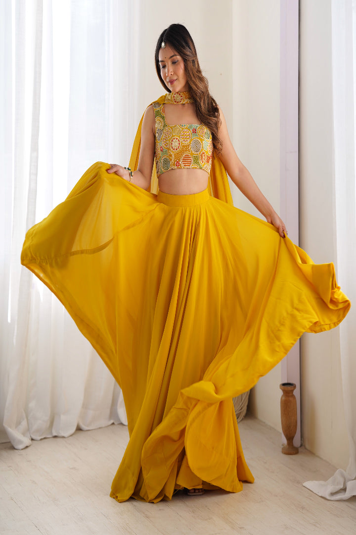 Woman in a yellow traditional outfit with a floral top and flowing skirt, standing against a white curtain background.