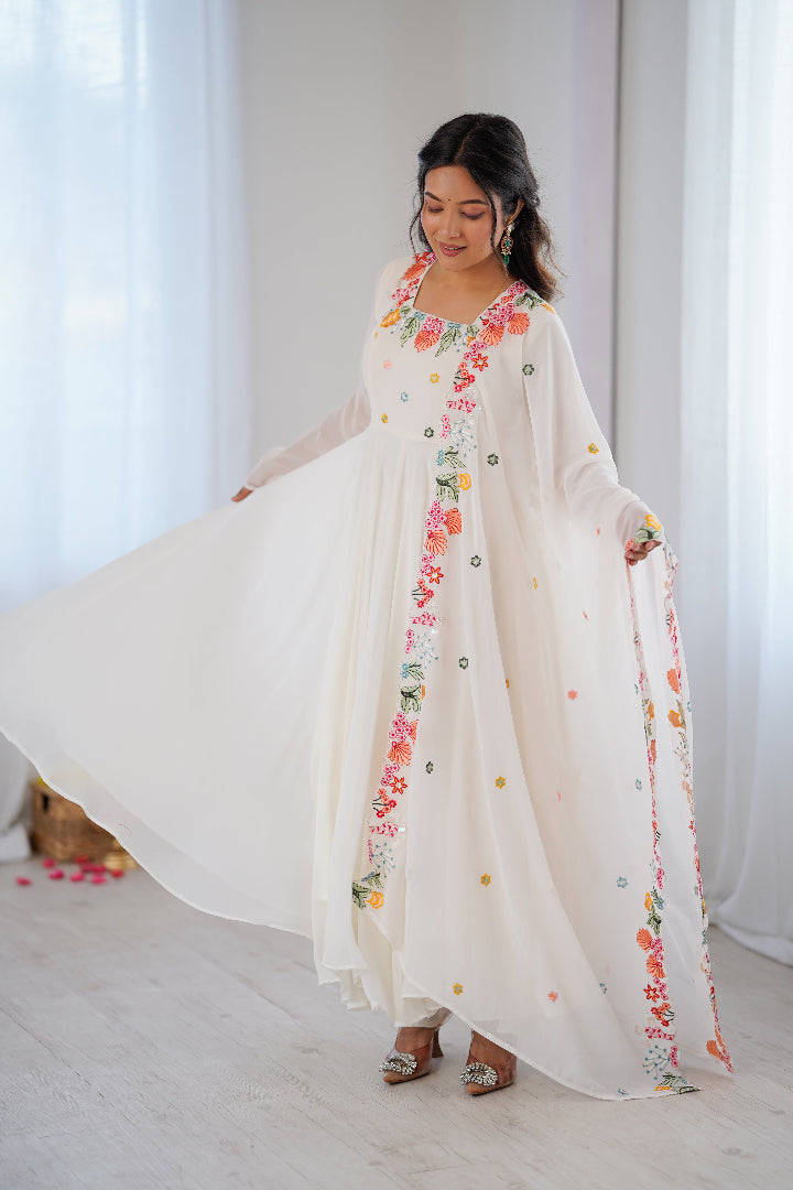 A woman modeling a long white caftan dress with colorful floral embroidery and dotted patterns, holding out the fabric to show its flow in a bright indoor setting.