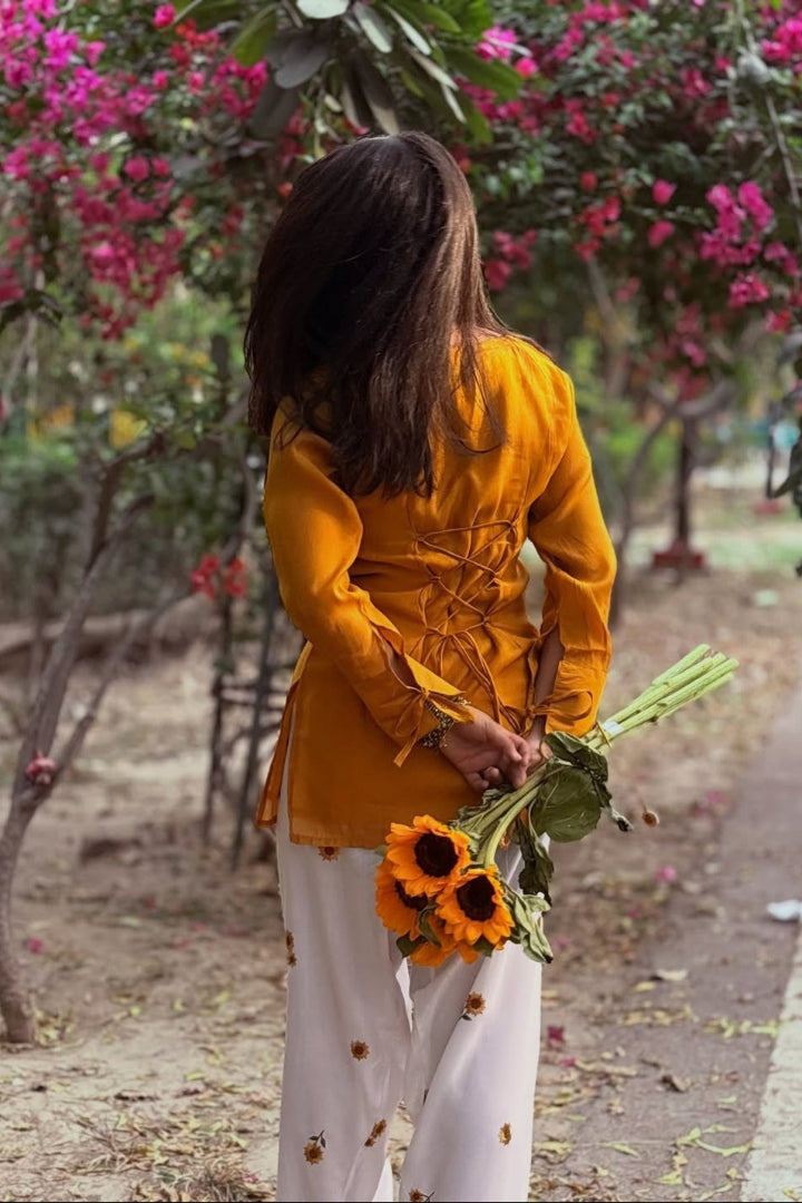 Person in a yellow top and white pants holding sunflowers in a garden with pink flowers.