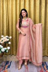 A woman models a peach-pink traditional Indian salwar kameez suit with a matching dupatta, set against a gold curtain backdrop.