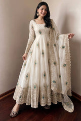 A woman modeling a long cream-colored traditional dress with intricate gold embroidery and green peacock feather motifs, paired with a matching sheer dupatta, in an indoor setting with a beige wall and dark wood floor.