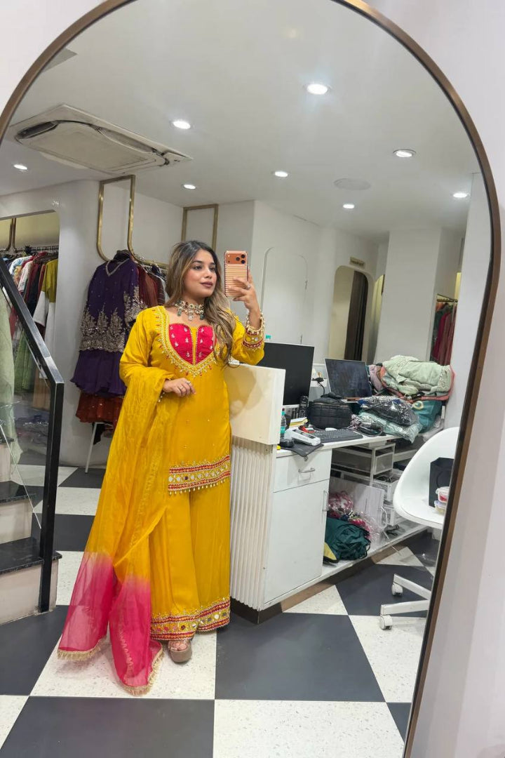 A woman takes a mirror selfie in a clothing boutique wearing a bright yellow traditional outfit with a pink ombre dupatta and red embroidered details on the bodice.