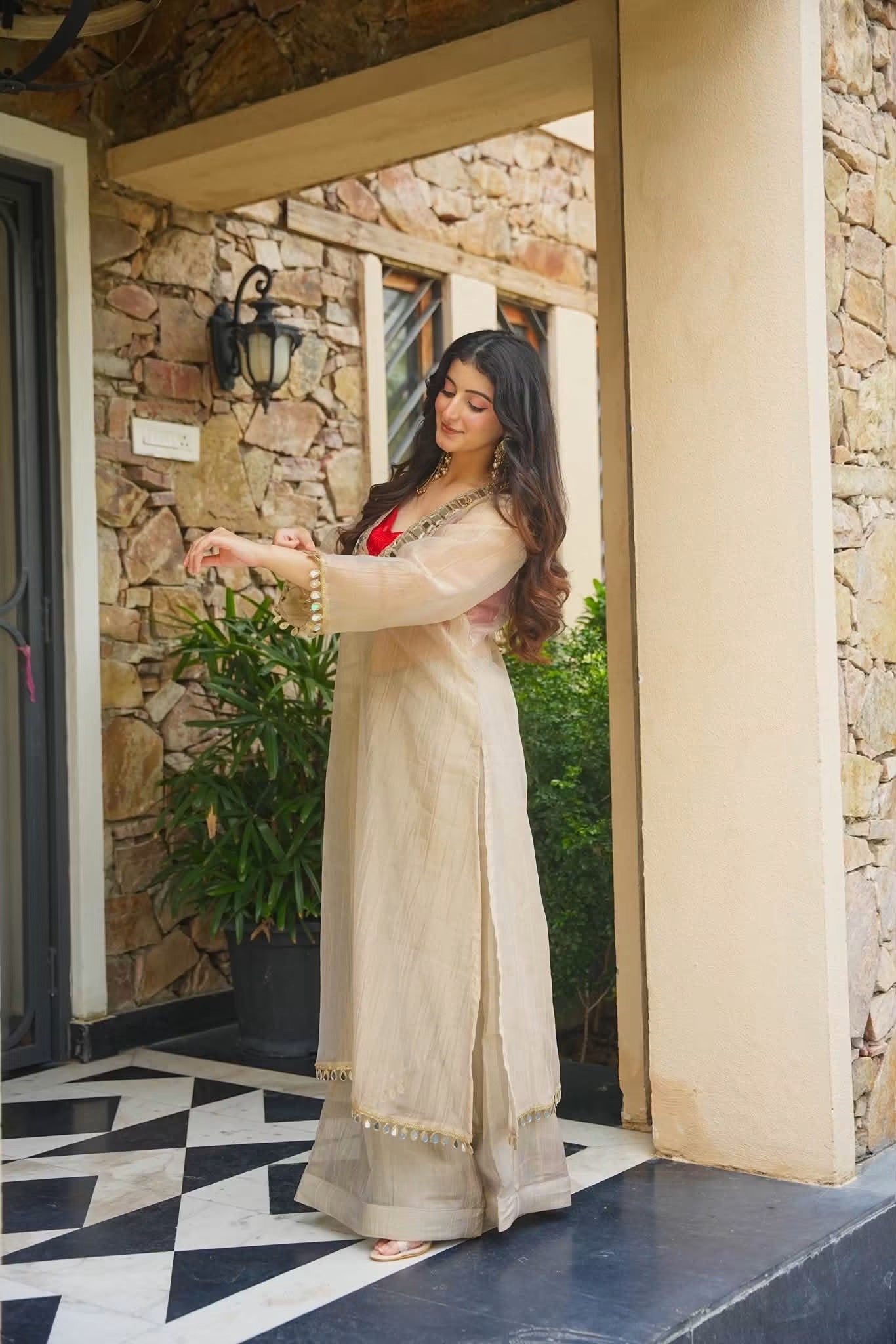 A woman wearing a beige long kurta with gold embroidery and a red inner dress, standing on a black and white checkered porch in front of a stone wall.
