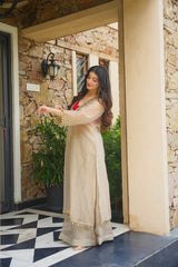 A woman wearing a beige long kurta with gold embroidery and a red inner dress, standing on a black and white checkered porch in front of a stone wall.