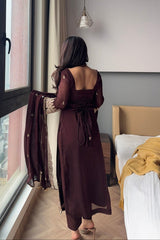 Woman in a long brown dress standing by a window in a bedroom.