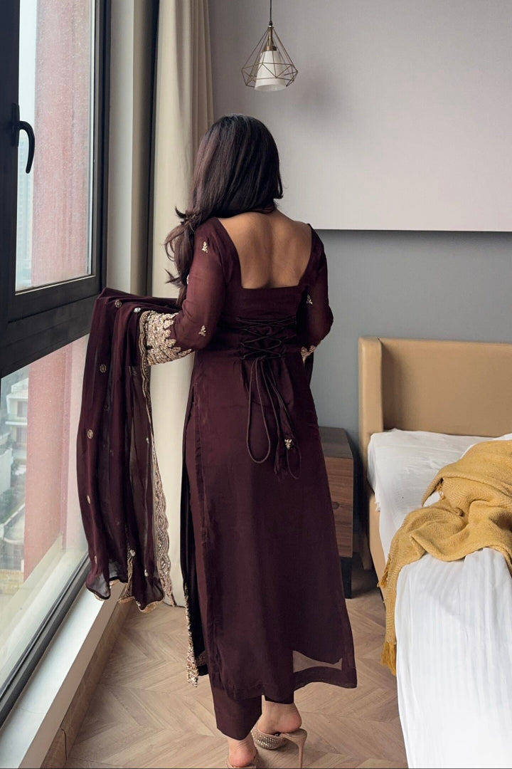 Woman in a long brown dress standing by a window in a bedroom.