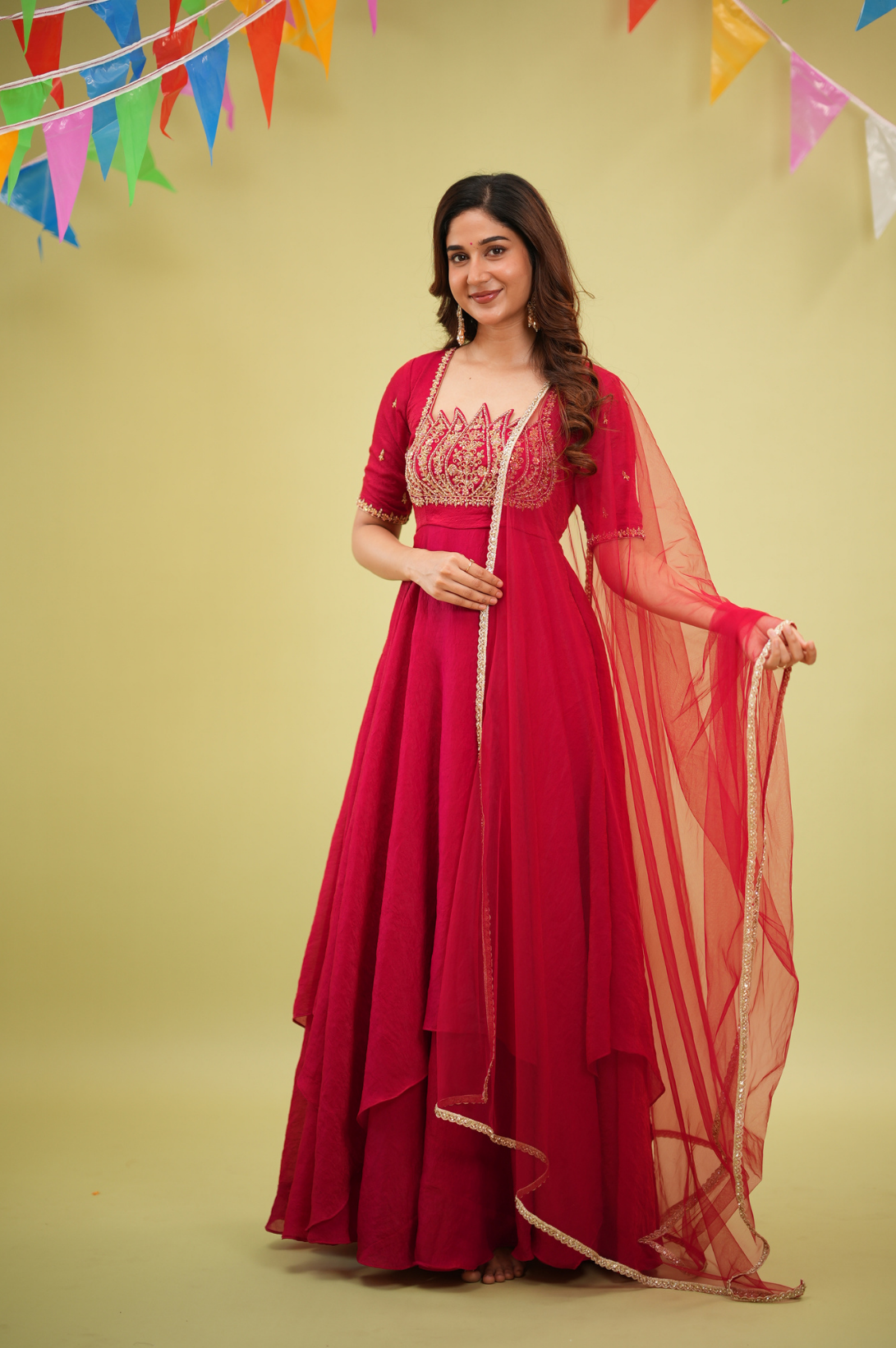 A woman models a deep red Anarkali gown with gold embroidery on the bodice, paired with a matching sheer red dupatta, set against a solid yellow background with colorful bunting.