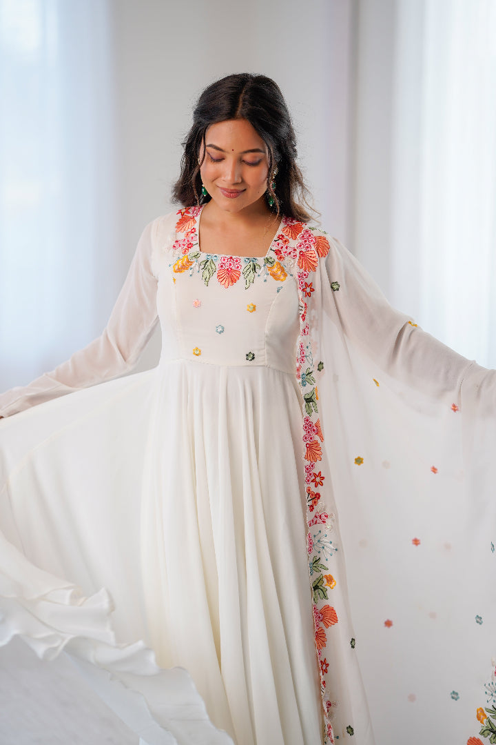 A woman wearing a long white embroidered dress with sheer sleeves and a matching embroidered dupatta, holding out the sides of the dress to show the full flow of the fabric in a bright indoor setting.