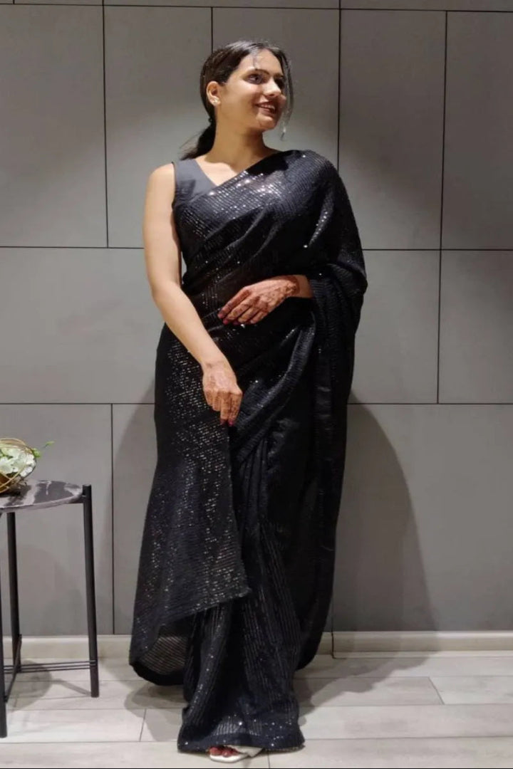 Woman in a black saree standing against a gray tiled wall.