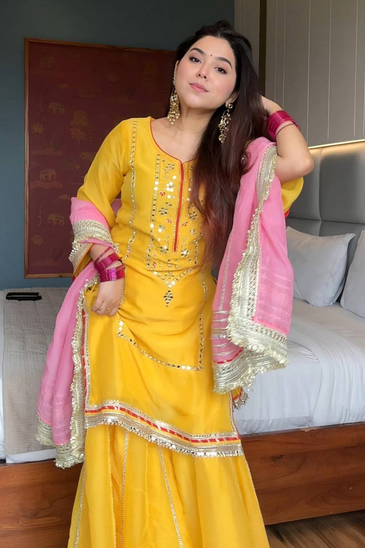 Woman in a yellow traditional outfit with pink dupatta in a room.