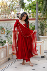 Woman in a red traditional outfit standing in an outdoor setting with plants and architecture.