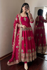A woman takes a mirror selfie wearing a bright magenta pink traditional Indian salwar kameez suit with intricate gold embroidery and green accents. The set includes a long kurta, matching trousers, and a dupatta.