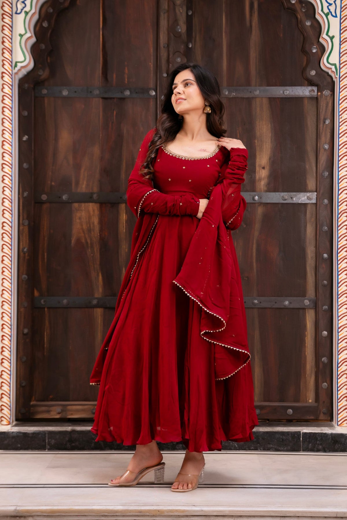 Woman in a red traditional outfit standing in front of a wooden door with decorative arches.