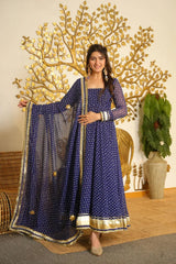 A woman models a navy blue Anarkali gown with gold and white polka dots, featuring a wide gold-trimmed border and holding out a matching sheer dupatta. She is standing in front of a decorative gold tree wall piece and a potted plant.