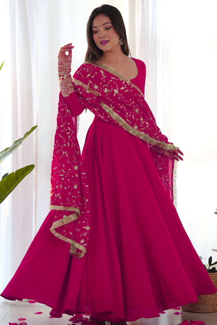 Woman wearing a pink saree with gold details against a white curtain background