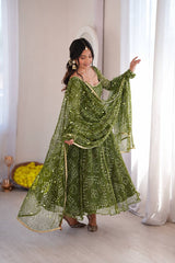 A woman modeling a green traditional Indian outfit with white printed motifs and a matching dupatta, standing in a bright room with white curtains.