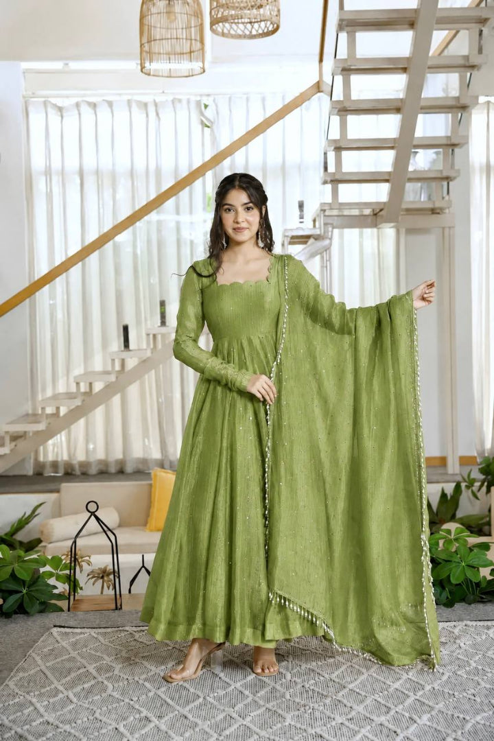 A woman models a sage green traditional Indian outfit consisting of a long-sleeved kurta, matching skirt, and a draped dupatta, set in a bright, modern home interior with a staircase in the background.
