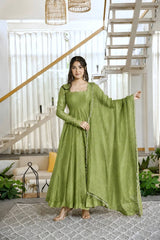 A woman models a sage green traditional Indian outfit consisting of a long-sleeved kurta, matching skirt, and a draped dupatta, set in a bright, modern home interior with a staircase in the background.