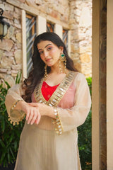 A woman wearing a traditional Indian outfit with a beige sheer kurta, gold coin embellishments, a pink embroidered blouse, and a red inner layer, standing outdoors in front of a stone wall.