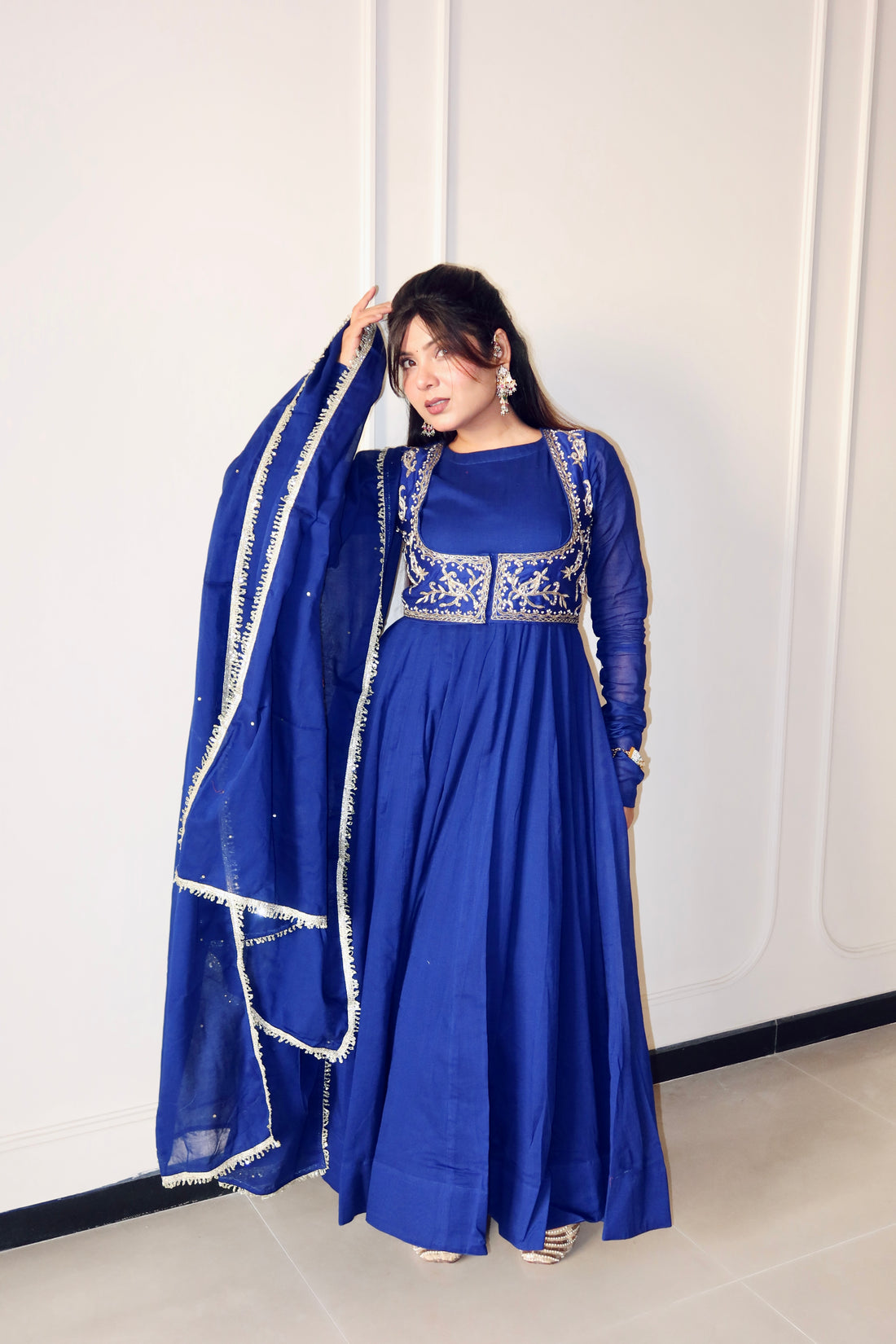 A woman models a royal blue Anarkali gown with silver embroidery on the bodice and sleeves, paired with a matching sheer dupatta featuring white border trim, set against a white paneled wall.