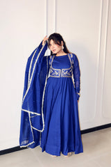 A woman models a royal blue Anarkali gown with silver embroidery on the bodice and sleeves, paired with a matching sheer dupatta featuring white border trim, set against a white paneled wall.