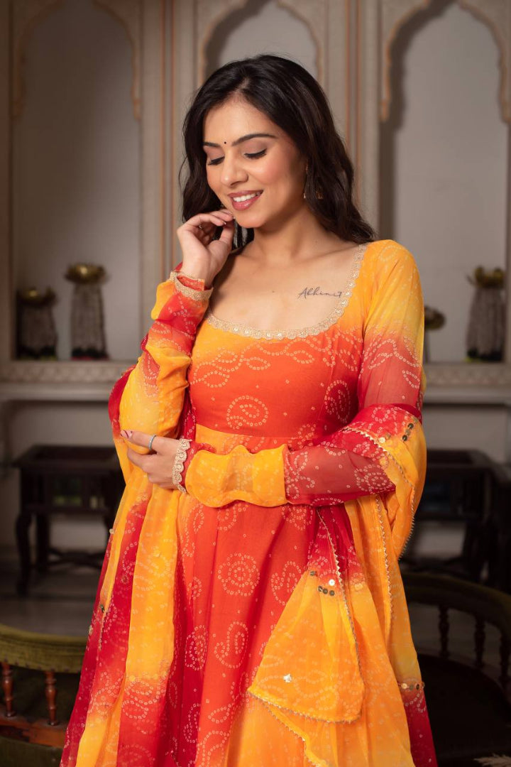 Woman wearing a traditional orange and yellow outfit with intricate patterns, standing in an indoor setting.