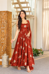 A woman modeling a rust-orange floor-length Anarkali gown with gold floral prints and a matching dupatta in an indoor setting with a staircase and wooden screen.