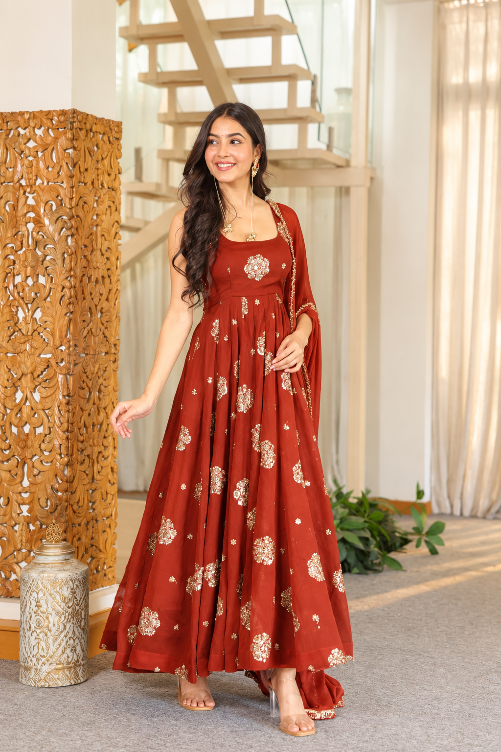 A woman modeling a rust-orange floor-length Anarkali gown with gold floral prints and a matching dupatta in an indoor setting with a staircase and wooden screen.