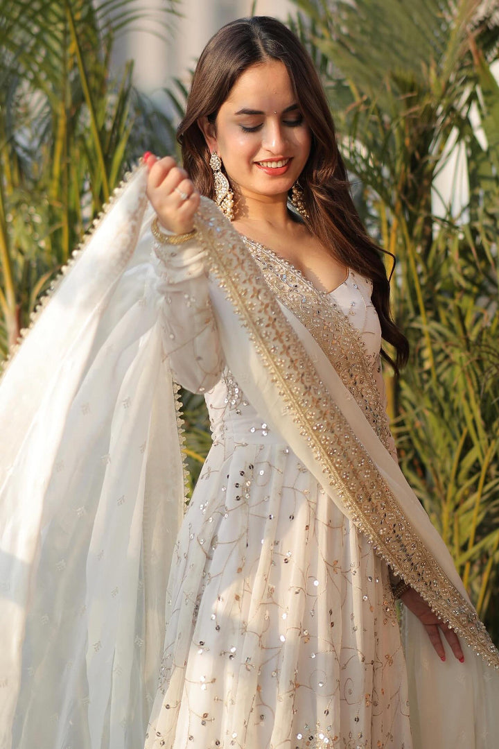 Woman in a white and gold traditional outfit with a dupatta, standing outdoors with greenery in the background.