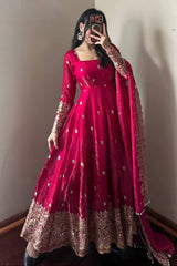Person wearing a vibrant pink traditional outfit with gold embroidery against a plain wall.