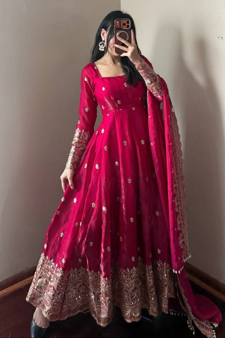 Person wearing a vibrant pink traditional outfit with gold embroidery against a plain wall.