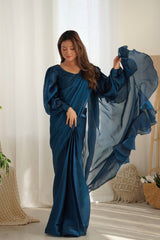 Woman in a blue saree standing indoors with white curtains in the background