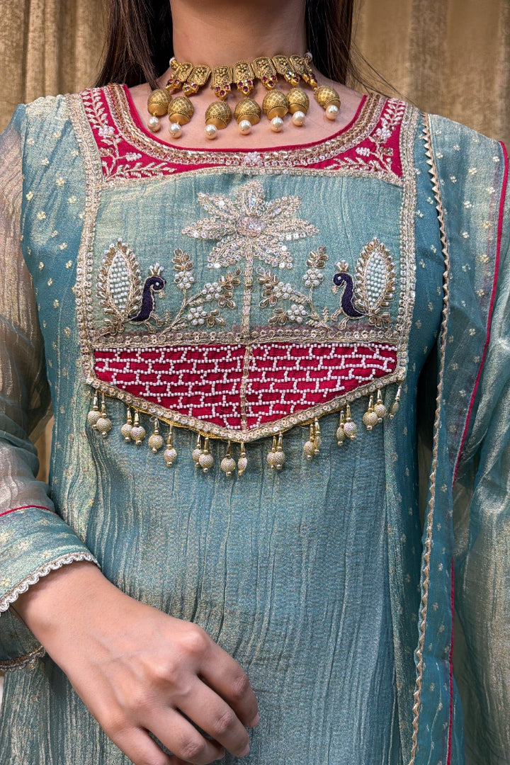 A close-up of a light blue-green embroidered traditional outfit with a red and gold embroidered chest panel, peacock motifs, bead tassels, and a matching sheer dupatta, worn by a model.