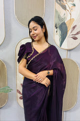 Woman in a purple saree standing against a decorative wall with floral patterns.