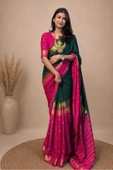 A woman modeling a dark green and magenta pink saree with gold zari embroidery, standing in front of a beige studio backdrop with a decorative vase of dried grass on the left.