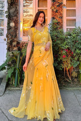 Woman in a yellow saree standing outdoors with plants and a building in the background