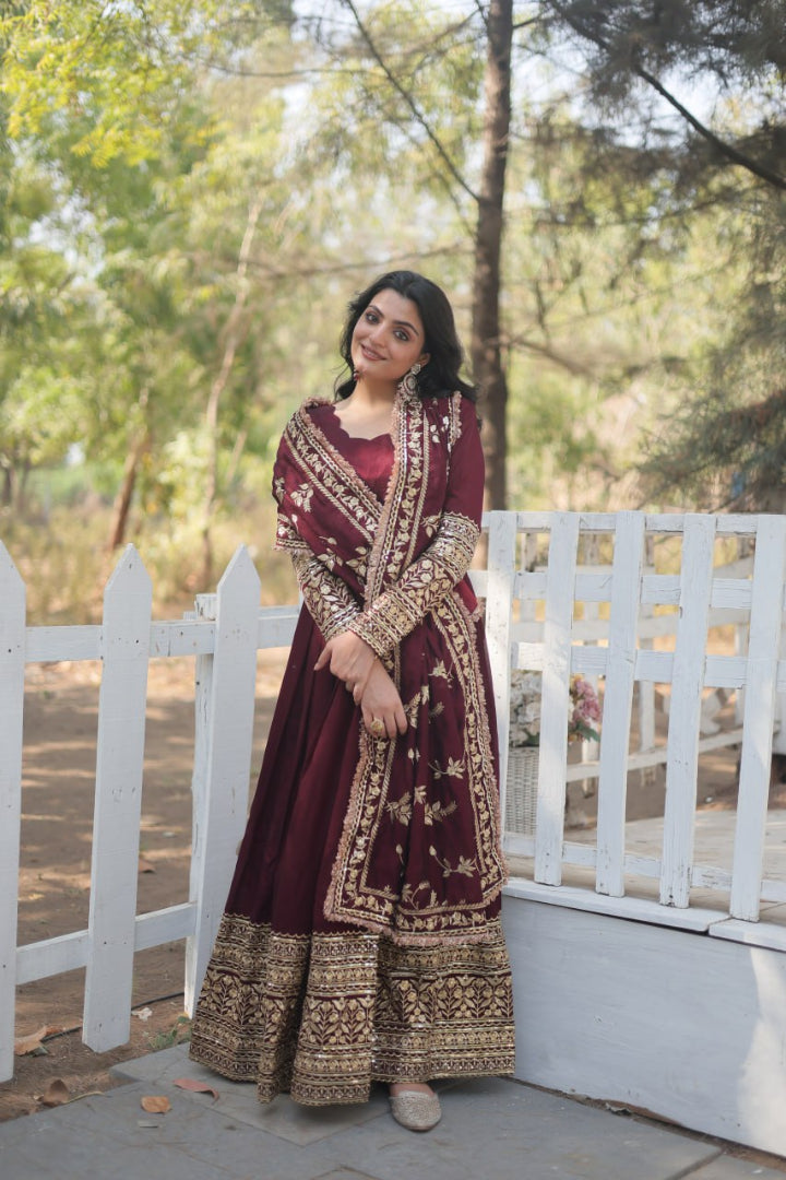 Woman in a maroon traditional outfit with gold embroidery standing outdoors near a white fence.