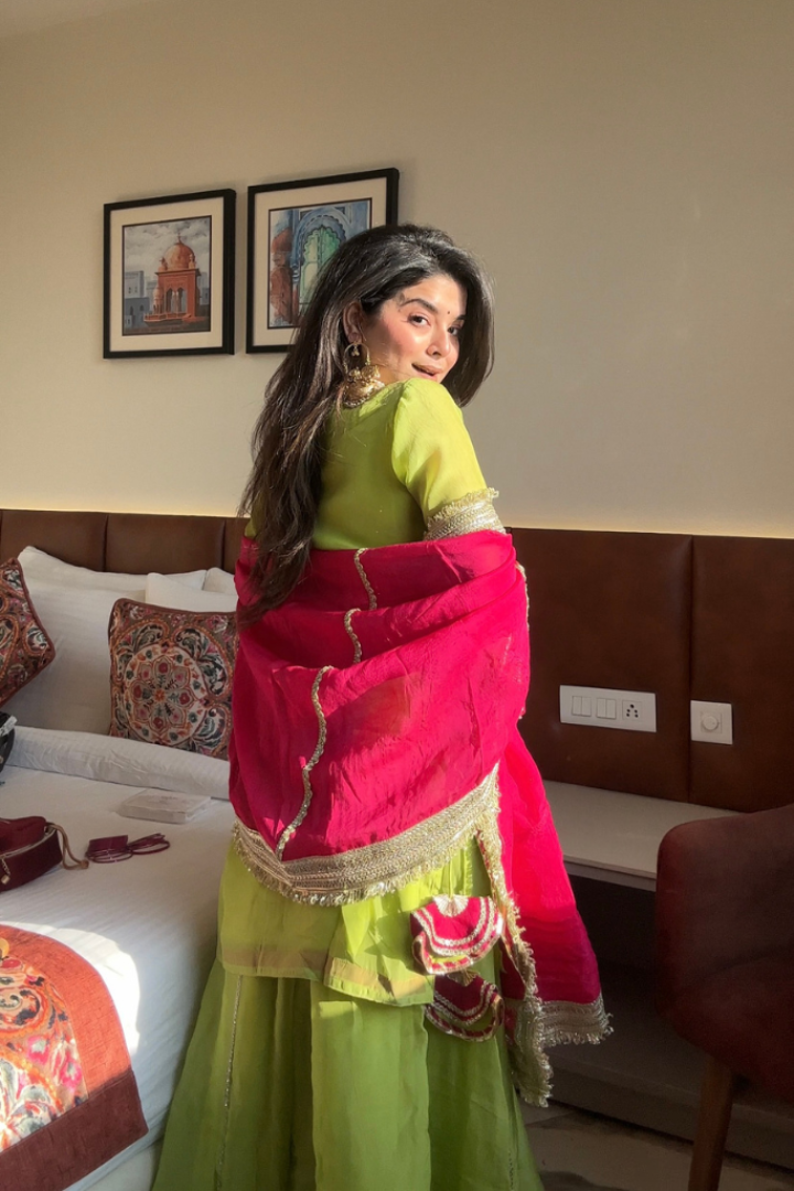 Woman in a green and pink traditional outfit standing in a room with a bed and wall art.