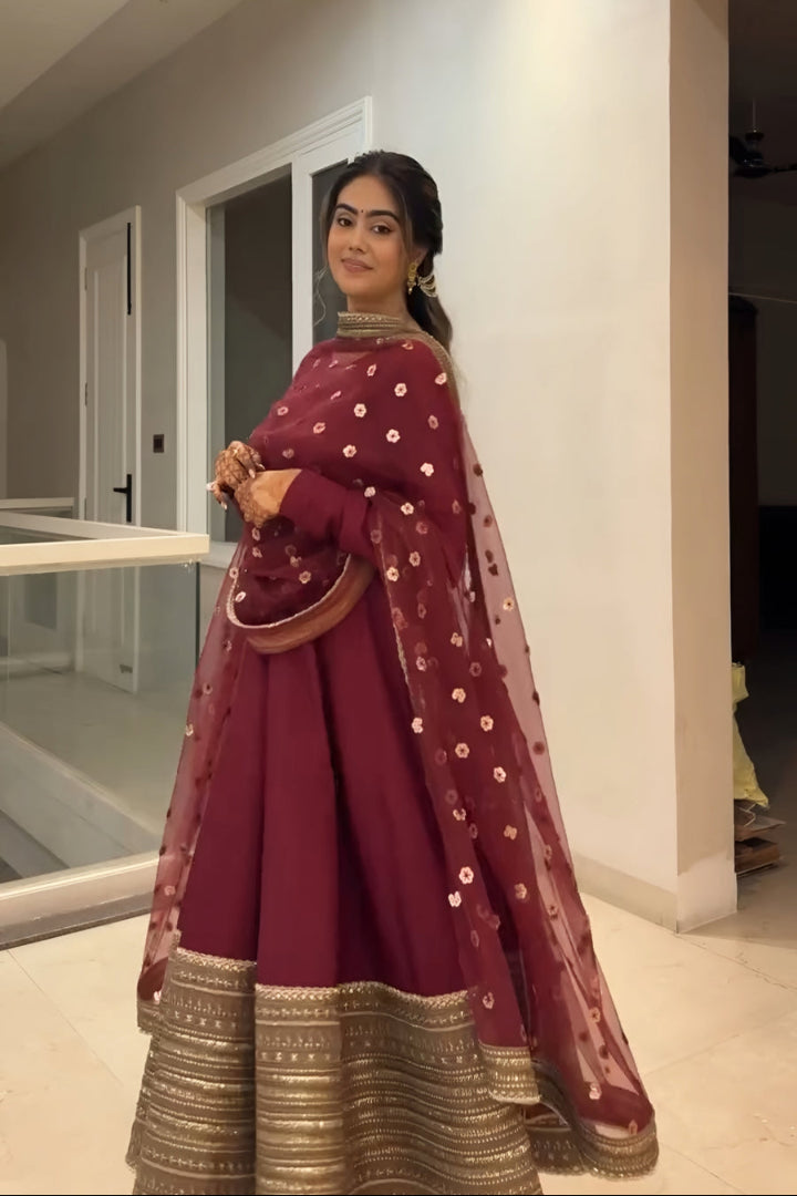 Woman in a maroon traditional outfit with gold details standing indoors.