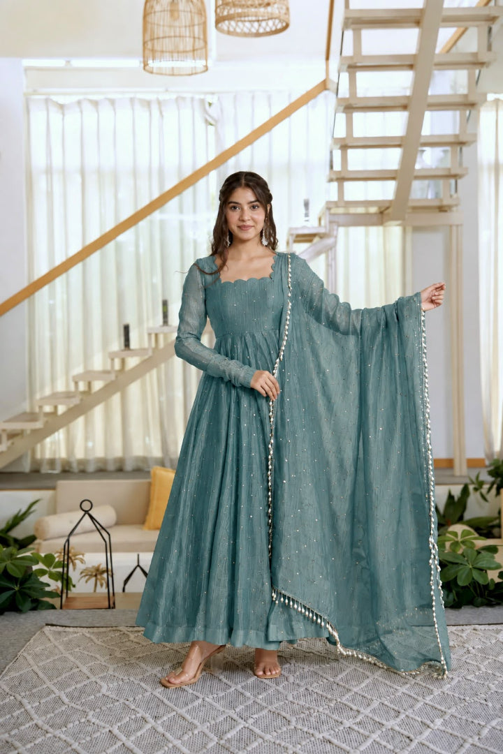 A woman models a dusty teal green ethnic salwar kameez set with a matching dupatta in a bright, modern living room with a staircase in the background.