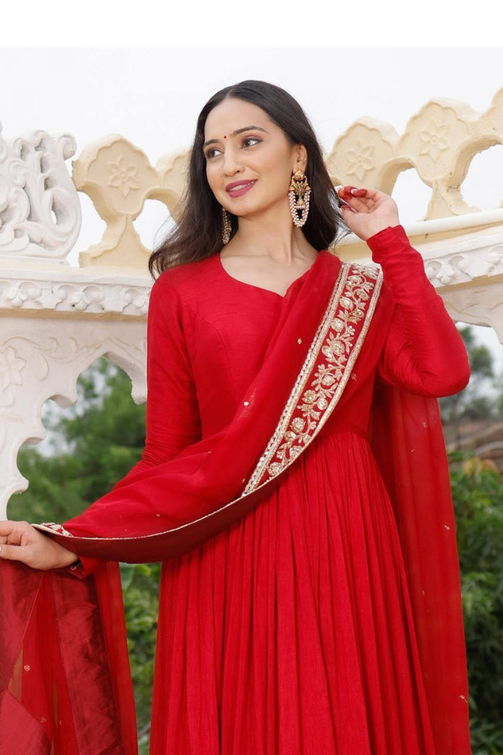 Woman in a red traditional outfit with gold details, standing in front of decorative stone carvings.