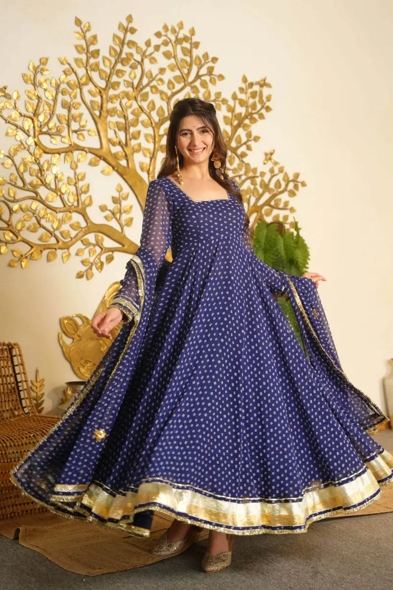A woman modeling a navy blue Anarkali gown with white polka dots and a gold border, standing in front of a decorative gold tree backdrop.